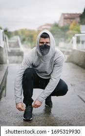 Sporty Man Wearing Face Mask For Protection Against Coronavirus And Pollution During Winter Running And Outdoor Fitness Workout Under The Rain. Sportsman Lacing Sport Shoes On Urban Stairs.