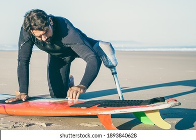 Sporty man in wetsuit wearing artificial limb, waxing surfboard on sand on ocean beach. Artificial limb and active lifestyle concept