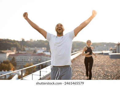 Sporty man in white t-shirt wins running competition from slim woman in black top during morning warm-up on roof of modern building on urban background. Healthy lifestyle concept. - Powered by Shutterstock