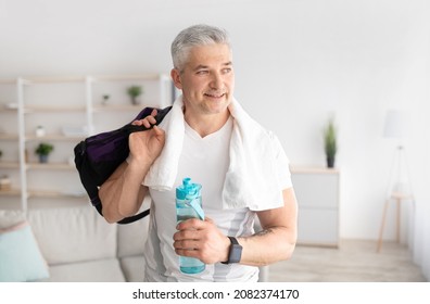 Sporty mature man holding bottle of water and sports bag, posing after domestic training and smiling, free space. Happy senior man keeping hydrated during home workout