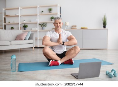 Sporty Mature Man Practicing Yoga Online With Instructor, Sitting In Lotus Position In Front Of Laptop On Mat In Living Room Interior, Looking And Smiling At Camera