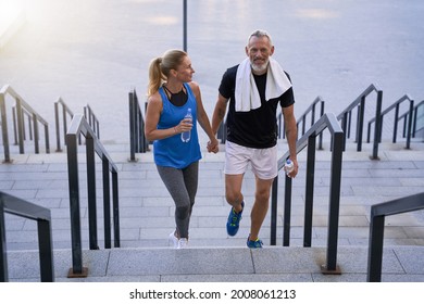 Sporty Middle Aged Couple In Love, Man And Woman In Sportswear Looking Happy, Holding Hands While Walking Up The Stairs After Training Together Outdoors