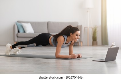 Sporty Millennial Woman Planking In Front Of Laptop, Living Room Interior, Side View, Copy Space. Healthy Young Lady In Sportswear Exercising At Home, Watching Online Fitness Class