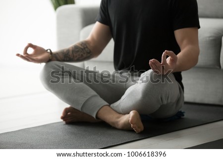 Sporty mindful man with tattoo meditating alone at home, peaceful calm hipster fit guy practicing yoga in lotus pose indoors holding hands in mudra, freedom and calmness concept, close up view