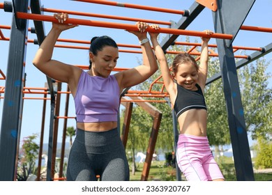 Sporty mother and daughter are bonding through doing sports together at an outdoor gym. Little girl hanging on the bar, learning how to do pull ups with her mom in the park. Close up, background. - Powered by Shutterstock