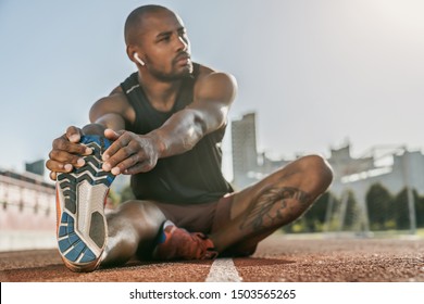 Sporty muscular african male athlete in earphones stretching legs while sitting at the stadium race track - Powered by Shutterstock