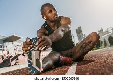 Sporty muscular african male athlete in earphones looking away, stretching legs while sitting at the stadium race track