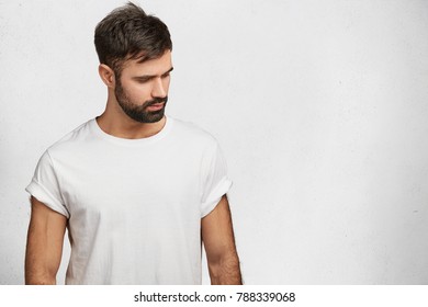 Sporty muscular man with trnedy hairdo, beard and mustache, wears white t shirt, looks with dreamy expression down, isolated over white concrete wall with copy space for your advrtisment or hearder