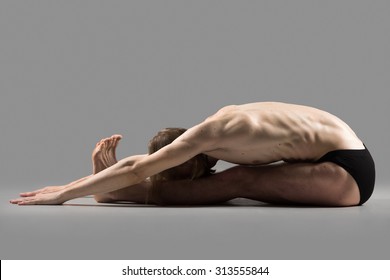 Sporty Muscular Young Yogi Man Sitting In Deep Paschimothanasana Pose, Seated Forward Bend Posture, Studio Shot On Dark Background, Profile View, Full Length