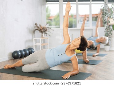 Sporty old woman practicing side plank pose of yoga on black rug during fitness classes - Powered by Shutterstock