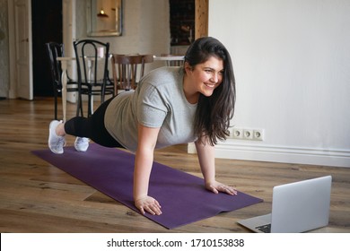 Sporty plus size woman in sportswear working out at home, doing plank on yoga mat in front of open laptop, repeating instructions by professional fitness trainer watching online video tutorial