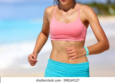 Sporty Running Woman Midsection Closeup Wearing A Pink Sports Bra And Smart Watch Activity Tracker Bracelet Band On Wrist. Fit Active Girl Runner On Summer Beach Exercising Cardio For Weight Loss.