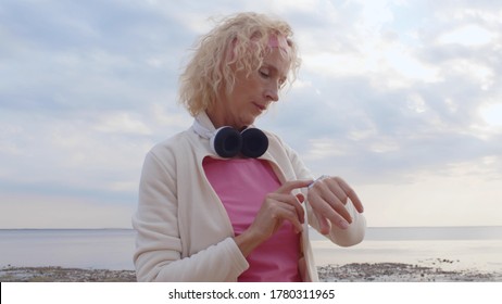 Sporty senior woman checking miles on smartwatch after running on beach. Side view of active mature female using smartwatch checking heartbeat after jogging on sea shore in morning - Powered by Shutterstock