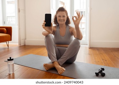 Sporty Senior Woman Holding Blank Smartphone And Showing Ok Gesture, Active Elderly Female Recommending New Fitness Tracker App, Sitting On Yoga Mat In Living Room Interior, Mockup - Powered by Shutterstock