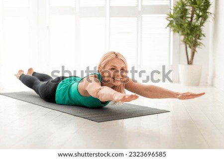 Sporty Senior Woman Making Superman Exercise While Training At Home, Smiling Mature Lady Doing Back Bending While Lying On Fitness Mat In Light Living Room, Raising Arms And Legs, Free Space