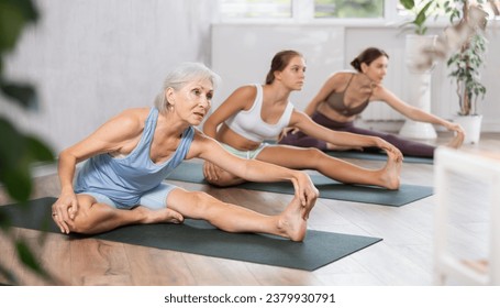 Sporty senior woman practicing yoga with group in modern studio, performing Janu Sirsasana seated asana to stretch hamstrings, back muscles, and spine - Powered by Shutterstock