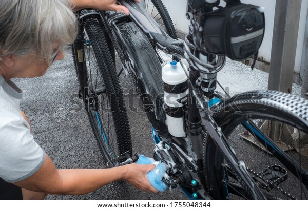 A sporty senior woman washes her bicycle with soap\
and sponge and takes care of the details. Service station with high\
pressure pump