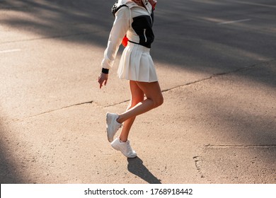Sporty slender woman in stylish skirt in sports jacket in fashion white sneakers. Modern girl walks on asphalt in city at sunset. Closeup of female sexy legs in sportswear and stylish shoes.