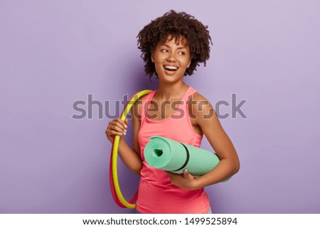 Sporty slim lady with healthy dark skin, Afro hairstyle, exercises with hula hoop, carries rolled up karemat, dressed in pink vest, has toothy smile, poses indoor. Healthy lifestyle, gymnastic concept