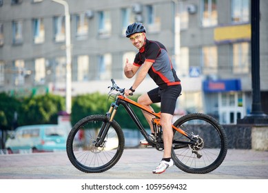 Sporty Smiling Guy Biker In Cycling Clothes And Helmet On Bicycle, Looking To The Camera, Showing Thumbs Up. Man Resting And Relaxing After Work, Training On Bicycle On City Streets. Healthy Lifestyle