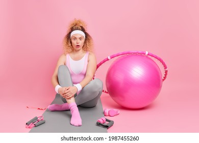 Sporty Surprised Woman Dressed In Activewear Poses On Fitness Mat Wears Headband Wristband Exercises With Fitball Hula Hoop Trains Pilates At Gym Poses Against Pink Background. Athletic Female Model