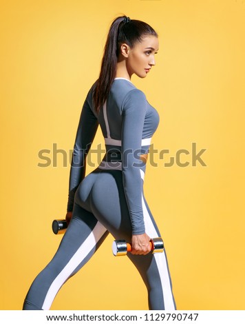 Sporty woman does the exercises with dumbbells. Photo of muscular woman in fashionable sportswear on yellow background. Strength and motivation.