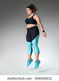 Sporty Woman Doing Jump After Exercise Squats. Photo Of Model With Curvy Figure In Fashionable Sportswear On Grey Background. Sports Motivation And Healthy Lifestyle