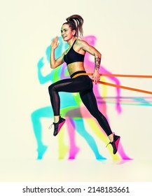Sporty woman doing jumping exercise with resistance band. Photo of muscular woman in black sportswear on white background with effect of rgb colors shadows. Dynamic movement. Side view