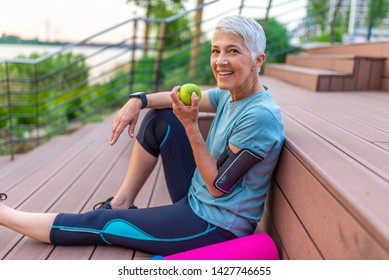 Sporty woman eating apple. Beautiful woman with gray hair in the early sixties relaxing after sport training. Healthy Age. Mature athletic woman eating an apple after sports training  - Shutterstock ID 1427746655