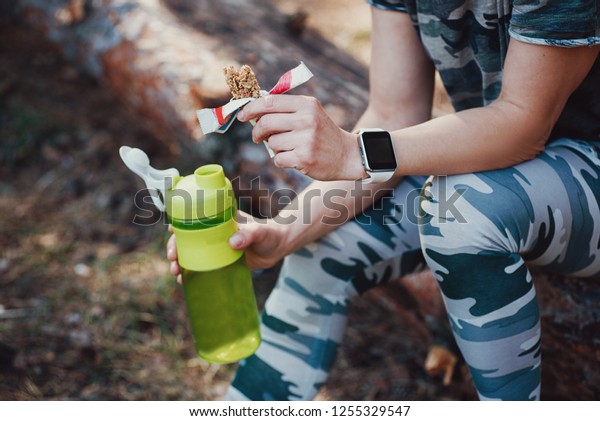 Sporty Woman\
Eating Protein Bar and drinking an amino acid cocktail. Woman in\
military-colored clothing in\
forest