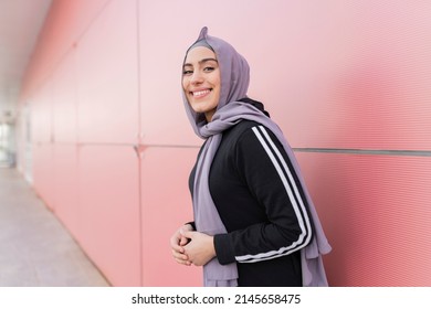 Sporty woman in hijab ready to run, on a red wall looking to the side with a smile on her face - Powered by Shutterstock