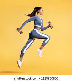 Sporty woman jumping with dumbbells. Photo of active woman in sportswear on yellow background. Dynamic movement. Side view. Sport and healthy lifestyle