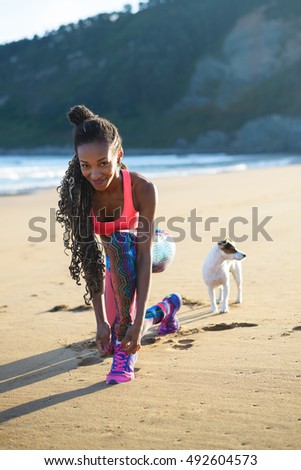 Sporty woman lacing sport footwear before running and exercising at the beach with her dog. Black beautiful runner training outdoor.