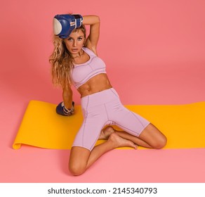 Sporty Woman Posing Sitting On Mat With Boxing Gloves On Pink Background