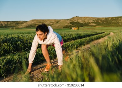 Sporty Woman Ready For Cross Country Running On Rural Track Surrounded By Field. Athlete Training And Exercising Outdoor At Sunset Or Morning.