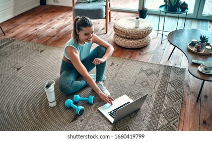 Спортивная женщина в спортивной одежде сидит на полу с гантелями и протеиновым коктейлем или бутылкой воды и использует ноутбук дома в гостиной. Концепция спорта и отдыха.