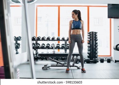 Sporty Woman Standing With Arms Crossed In Fitness Gym In Full Size. Serious Concentrated Sporty Girl Is Examining Her Muscle And Body Looking Into The Mirror