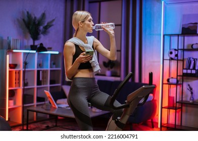 Sporty Woman Using Mobile Phone, Drinking Water While Riding On Exercise Bike At Home At Night Time. Beautiful Sporty Fit Female Cycling Stationary Bike And Watching Online Fitness Class On Smartphone