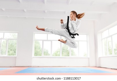 Sporty Woman In White Kimono With Black Belt Jumping And Performing Kick. Young Girl Confidently Doing Complex Technique Elements Karate With Serious Facial Expression. Healthy Lifestyle Concept.