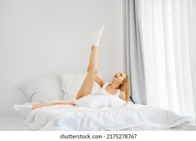 Sporty Woman In White Nightwear Having Fun In Bed, Stretching One Leg Up