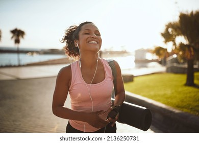Sporty young African woman laughing and listening to music with her cellphone while walking along a promenade carrying a yoga mat - Shutterstock ID 2136250731