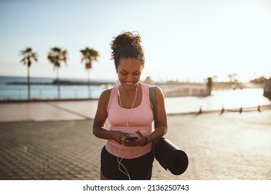 Sporty young African woman walking with a yoga mat along a promenade queueing up a music playlist on her cellphone    - Powered by Shutterstock