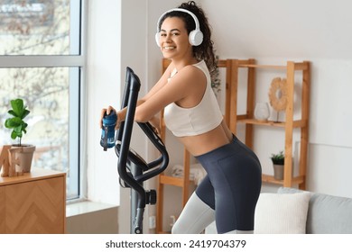 Sporty young African-American woman in headphones exercising on bike at home - Powered by Shutterstock