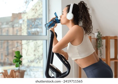 Sporty young African-American woman in headphones drinking water on exercising bike at home - Powered by Shutterstock