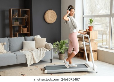 Sporty young African-American woman with headphones training on treadmill in living room - Powered by Shutterstock