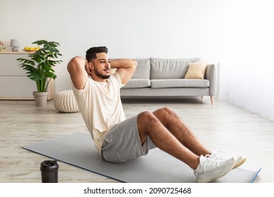 Sporty young Arab man doing abs exercises, working out at home, full length portrait. Athletic middle Eastern guy doing domestic training during covid pandemic, strengthening core muscles - Shutterstock ID 2090725468