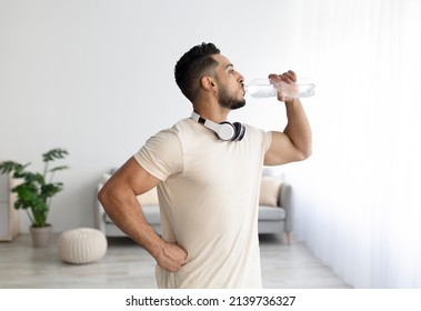 Sporty young Arab man with headphones drinking water after his workout at home. Fit Eastern guy staying hydrated during domestic sports training. Healthy lifestyle, wellness concept