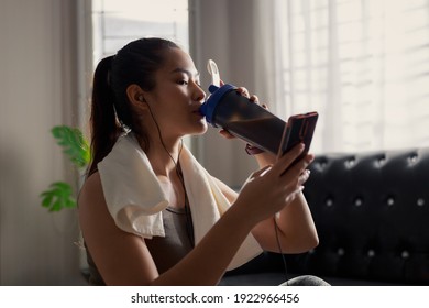 Sporty young Asian female enjoying healthy food and restoring energy after routine fitness workout at home. Fit woman sitting on sports mat with mobile phone and headphones.