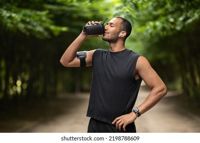 Sporty young black man drinking protein cocktail, training at public park, using wireless earbuds, listening to music while jogging by forest, copy space. Sports nutrition concept - Powered by Shutterstock