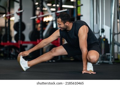 Sporty Young Black Man Stretching Leg Muscles While Training At Gym, Smiling African American Male Athlete Warming Up Before Workout In Modern Sport Club, Motivated Guy Exercising Indoors, Copy